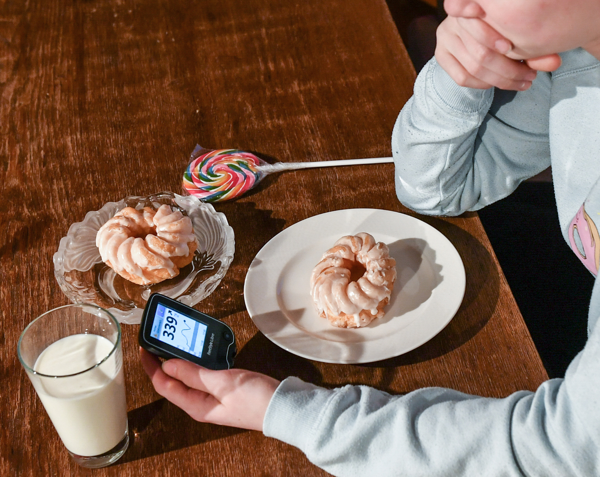 A girl is holding a Freestyle Libre blood glucose meter with far too high a blood glucose level next to plates of cake, milk and lollipops in her hands.