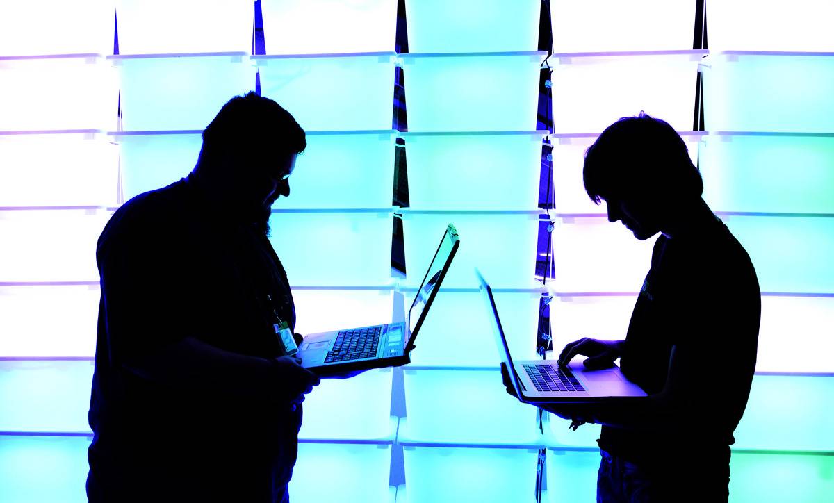 Two men browse through their laptops as light illuminates them from behind.